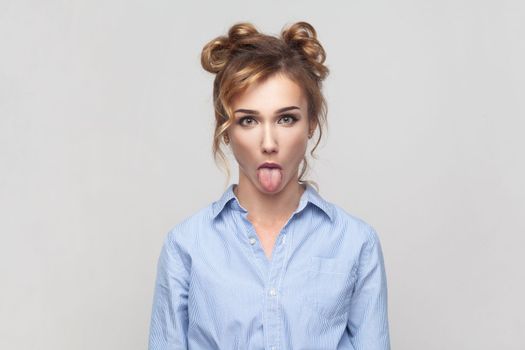 Portrait of naughty comic blonde woman sticking out tongue, misbehaves, demonstrating her disobedience and protest, has funny look, wearing blue shirt. Indoor studio shot isolated on gray background.