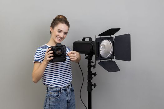 Portrait of smiling satisfied woman photographer photographing, holding photo camera and using illumination equipment, pointing to camera. Indoor studio shot isolated on gray background.