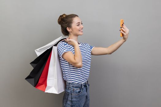 Side view portrait of woman wearing striped T-shirt broadcasting livestream and boasting her purchase, holding shopping, using smartphone. Indoor studio shot isolated on gray background.