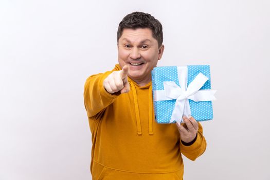 Portrait of satisfied middle aged handsome man standing, holding present box, pointing finger to camera, wearing urban style hoodie. Indoor studio shot isolated on white background.