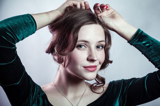 Beauty portrait of smiling delighted adorable beautiful woman with freckles wearing green dress standing holding her hair, looking at camera. Indoor studio shot isolated on gray background.