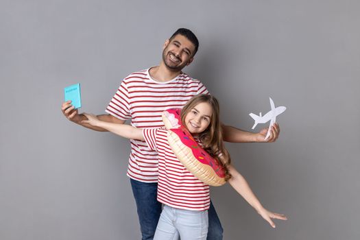 Portrait of excited smiling father and daughter in striped T-shirts holding passport and rubber ring, journey abroad, vacation to the sea. Indoor studio shot isolated on gray background.