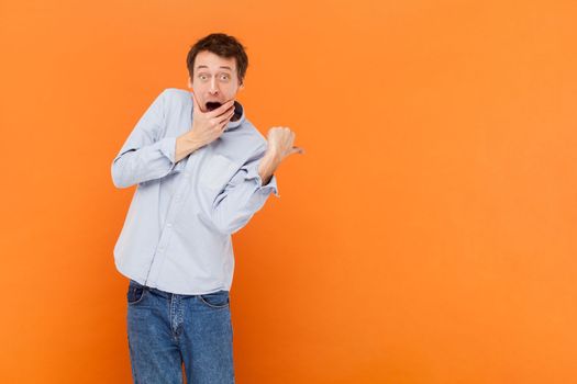 Man looks with jaw dropped out at camera, being horrified to see something, indicates with thumb aside, advertisement area, wearing light blue shirt. Indoor studio shot isolated on orange background.
