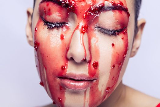 Beauty closeup portrait of calm beautiful young adult woman in a raspberry or strawberry jam flowing on her face, keeps eyes closed. Indoor studio shot isolated on gray background.