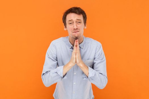 Portrait of hopeful handsome man standing with palms together, asking to forgive, pleading, looking at camera with pout lips, wearing light blue shirt. Indoor studio shot isolated on orange background
