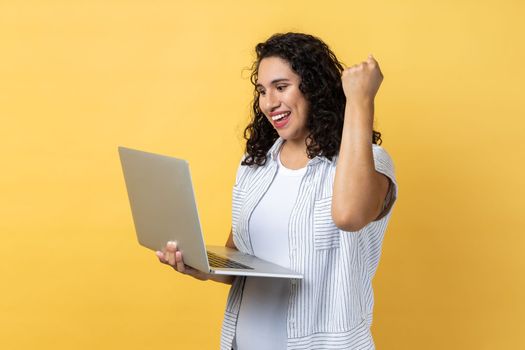 Portrait of extremely happy woman with dark wavy hair working on laptop and looking at display, clenched fist, celebrating her winning. Indoor studio shot isolated on yellow background.