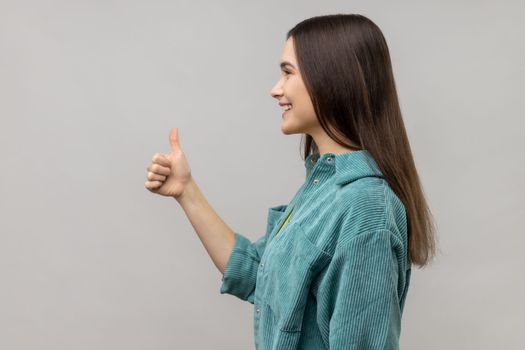 Side view of woman looking ahead with smile and showing thumb up, approval sign, satisfied with service, good feedback, wearing casual style jacket. Indoor studio shot isolated on gray background.