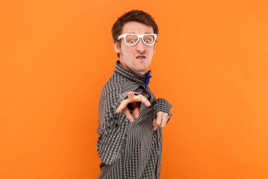 Portrait of funny crazy disabled man nerd pointing and looking at camera with clenched teeth, wearing shirt with blue bow tie and white glasses. Indoor studio shot isolated on orange background.