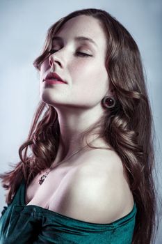 Beauty portrait of relaxed calm winsome woman with beautiful earrings wearing green dress, showing her bare shoulders, keeps eyes closed. Indoor studio shot isolated on gray background.