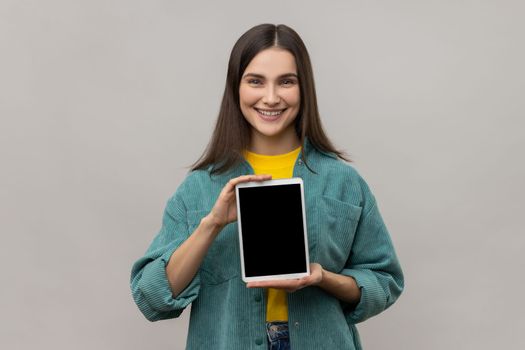 Portrait of positive delighted woman holding showing tablet, looking at camera with smile and happiness, wearing casual style jacket. Indoor studio shot isolated on gray background.