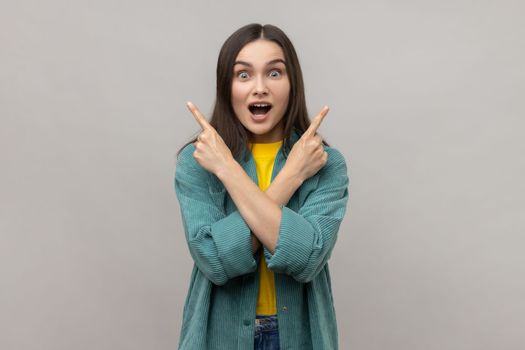 Portrait of surprised woman with dark hair standing with crossed hands and pointing to copy space on both sides, wearing casual style jacket. Indoor studio shot isolated on gray background.