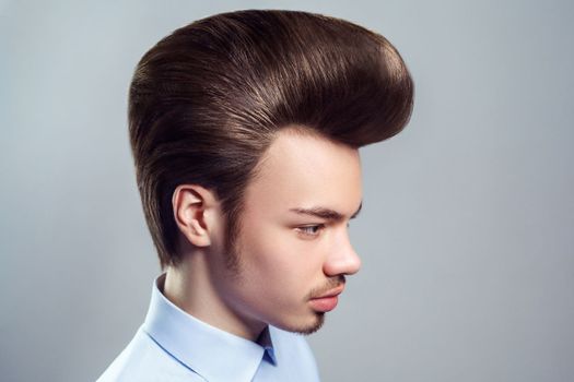 Side view portrait of young adult attractive man with mustache and beard with retro classic pompadour hairstyle, looking away with serious expression. Indoor studio shot isolated on gray background.