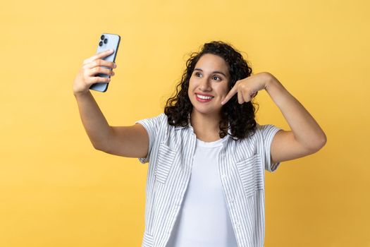 Portrait of joyful cheerful woman with dark wavy hair holding phone making selfie or broadcasting livestream, pointing down, asking to subscribe. Indoor studio shot isolated on yellow background.