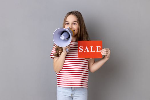 Little girl wearing striped T-shirt holding card with sale inscription and megaphone near mouth, loudly speaking, screaming, making announcement. Indoor studio shot isolated on gray background.