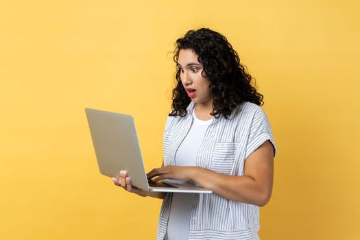 Portrait of shocked astonished woman with dark wavy hair working on laptop, looking at screen with big eyes and open mouth. Indoor studio shot isolated on yellow background.