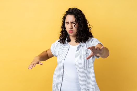 I can't see. Portrait of woman with dark wavy hair standing with closed eyes and outstretched hands, trying to find obstacles while walking blind. Indoor studio shot isolated on yellow background.