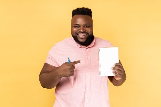 Write to-do list here into notepad. Portrait of man wearing pink shirt showing empty paper page, template mock up for ideas, wearing white shirt. Indoor studio shot isolated on yellow background.
