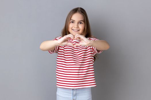 Portrait of little girl wearing striped T-shirt holding hands in shape of heart showing romantic gesture, love confession, valentines day celebration. Indoor studio shot isolated on gray background.