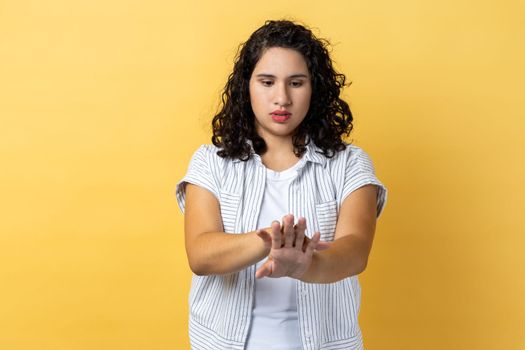 Portrait of beautiful young adult concentrated woman with dark wavy hair standing looking at her fingers, looks at her manicure. Indoor studio shot isolated on yellow background.
