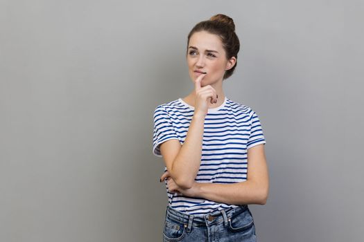 Portrait of woman wearing striped T-shirt pondering serious issues, looking with uncertain hesitant expression, making difficult choice. Indoor studio shot isolated on gray background.
