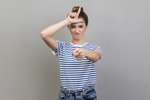 Portrait of woman wearing striped T-shirt expressing disrespect, showing L finger sign to camera, loser gesture, accusing for failure. Indoor studio shot isolated on gray background.