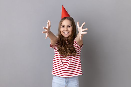 Portrait of pretty little girl wearing striped T-shirt and party cone on head giving free hugs with outstretched hands, welcoming inviting to embrace. Indoor studio shot isolated on gray background.