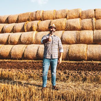 Happy farmer is standing beside bales of hay. He is satisfied because of successful harvesting.