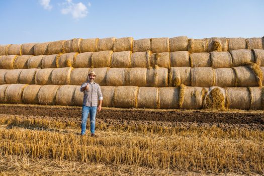 Happy farmer is standing beside bales of hay. He is satisfied because of successful harvesting.