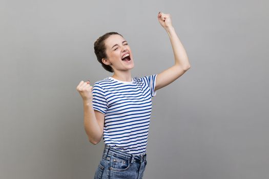 Portrait of happy satisfied woman wearing striped T-shirt showing yes gesture, screaming celebrating her victory, success, dreams comes true, euphoria. Indoor studio shot isolated on gray background.
