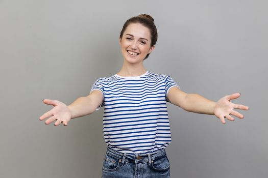 Please, take it. Portrait of woman wearing striped T-shirt standing with wide open hands as if sharing for free, giving charity and smiling. Indoor studio shot isolated on gray background.
