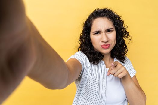 Beautiful young adult woman blogger with dark wavy hair making point of view photo or broadcasting livestream, pointing finger to camera, POV. Indoor studio shot isolated on yellow background.