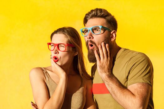 Portrait of amazed young adult couple in colorful glasses looking away with astonished facial expression, being shocked and surprised. Indoor studio shot isolated on yellow background.