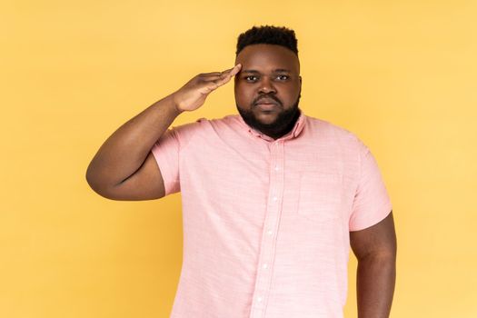 Yes sir. Portrait of serious man wearing pink shirt holding hand near head as soldier and saluting, looking attentive and confident. Indoor studio shot isolated on yellow background.