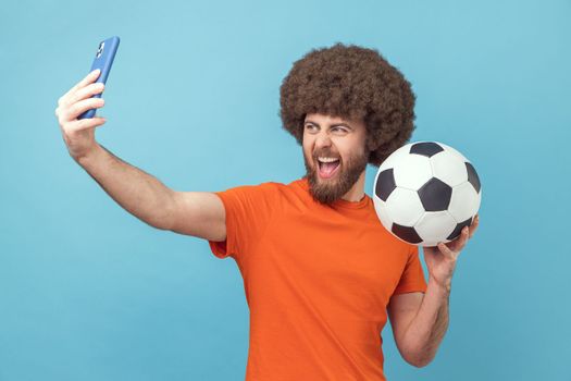 Portrait of excited man with Afro hairstyle wearing orange T-shirt standing with soccer ball and broadcasting livestream from football match. Indoor studio shot isolated on blue background.