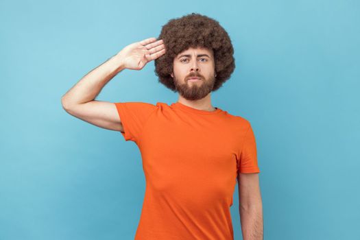 Portrait of man with Afro hairstyle wearing orange T-shirt saluting with respect as if soldier waiting order from commander, obeying discipline. Indoor studio shot isolated on blue background.