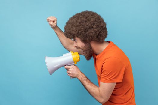 Side view of serious man with Afro hairstyle wearing orange T-shirt raised hands and holding megaphone, screaming in loud speaker, protesting. Indoor studio shot isolated on blue background.