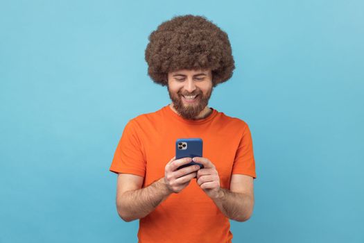 Portrait of positive man blogger with Afro hairstyle wearing orange T-shirt standing, using smartphone and watching video with happy smiling face. Indoor studio shot isolated on blue background.