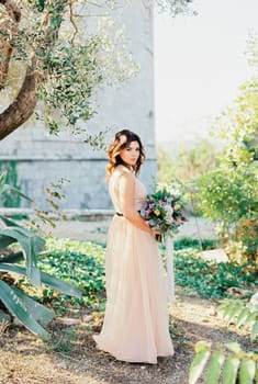 Bride with a bouquet stands half-turned near a green tree. High quality photo
