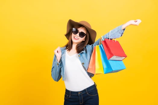 Asian happy portrait beautiful cute young woman teen smiling standing with sunglasses excited holding shopping bags multi color looking camera isolated, studio shot yellow background with copy space