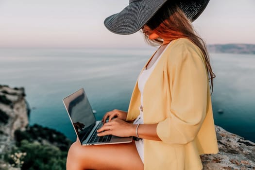 Successful business woman in yellow hat working on laptop by the sea. Pretty lady typing on computer at summer day outdoors. Freelance, travel and holidays concept.