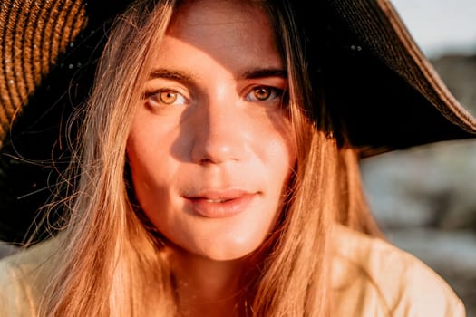 Portrait of happy young woman wearing summer black hat with large brim at beach on sunset. Closeup face of attractive girl with black straw hat. Happy young woman smiling and looking at camera at sea