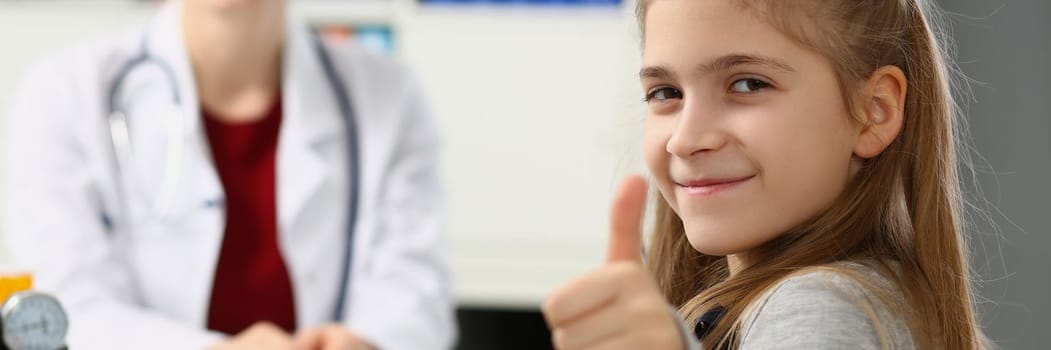 Beautiful little girl shows thumbs up. and doctor sits in background. Health insurance and examination of children