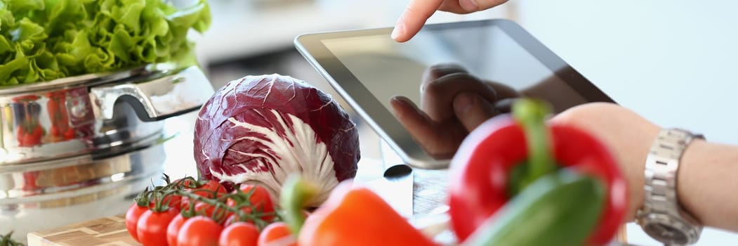 Person holding digital tablet and fresh vegetables in kitchen. Online application for shopping or food recipes and organic farm products