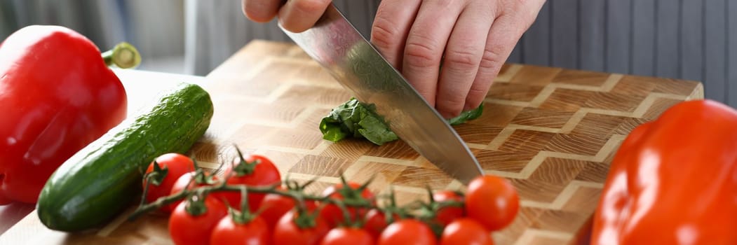 Chef is preparing salad of spinach cucumbers and tomatoes in kitchen. Closeup of chef hands cutting vegetables with knife on cutting board