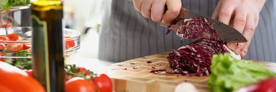 Male hands are chopping red cabbage on cutting board surrounded by various fresh vegetables on table. Closeup of male hands slicing fresh vegetables on cutting board