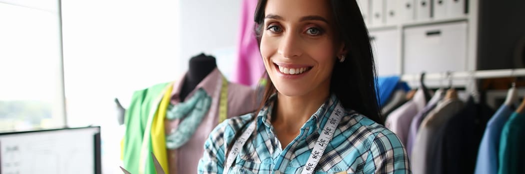 Young woman smiles and stands in tailor workshop with scissors in hand. Atelier services and tailoring concept