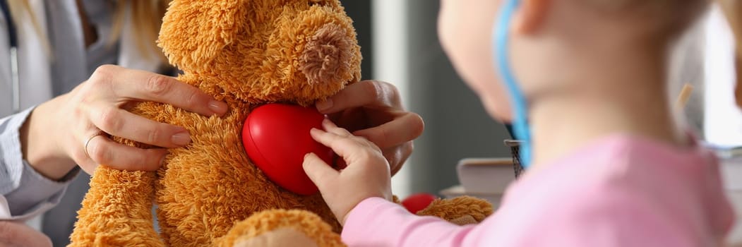 Little child girl plays with plush toy at doctor appointment. Child listens to soft toy with stethoscope and holds heart. Treatment of cardiac diseases in children
