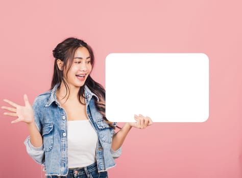 Happy Asian beautiful young woman smiling excited wear denims holding empty speech bubble sign, Portrait female posing show up for your idea looking at bubble, studio shot isolated on pink background