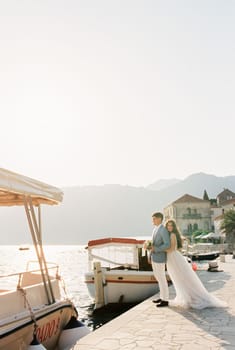 Bride hugs groom from behind on the pier near the moored boats. High quality photo