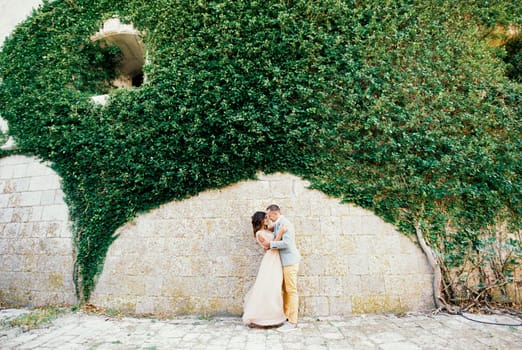 Groom hugs bride near the arch of green ivy on the stone wall. High quality photo